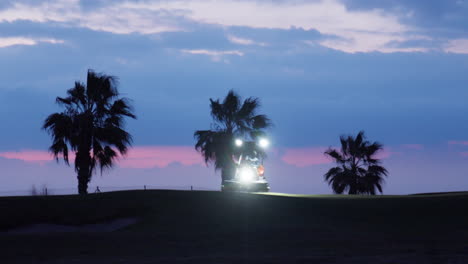Ein-Traktor-Mit-Scheinwerfern-Mäht-In-Der-Blauen-Stunde-Des-Frühen-Morgens-Das-Gras-Auf-Einem-Golfplatz,-Während-Der-Wind-Die-Silhouetten-Der-Palmen-Im-Hintergrund-Raschelt