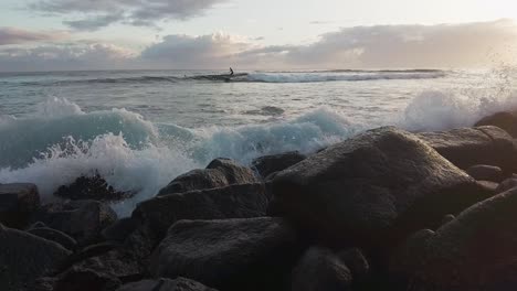 Plätschernde-Wellen-Treffen-Auf-Die-Küste,-Surfer-Im-Hintergrund,-Zeitlupe