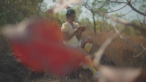 Blurly-shot-of-a-young-woman-through-leaf-sitting-on-a-rock-surrounded-by-trees-in-a-field