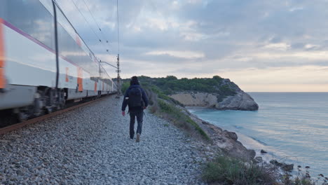 A-man-walks-along-train-tracks-as-a-passenger-train-rushes-by-nearby,-while-the-calm-sea-washes-down-the-cliff-side
