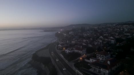 Aerial-view-of-cascais-city-in-Portugal,-drone-fly-above-coastline-with-some-cars-driving-and-small-waves-crashing-on-shore