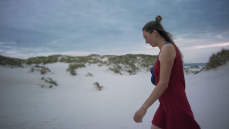 Young-woman-walks-at-a-white-sand-beach-wearing-a-red-dress
