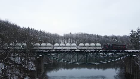 Tracking-drone-shot-of-cargo-train-crossing-a-bridge-over-a-lake-in-the-middle-of-a-snowy-forest-in-the-Kashubian-District,-Poland