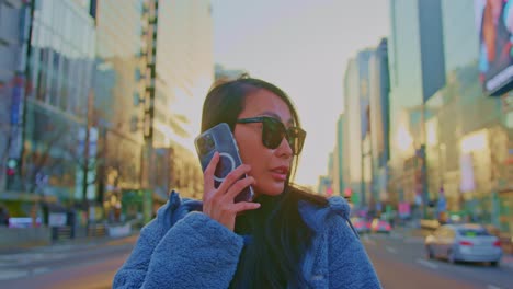 beautiful-Asian-oriental-female-girl-woman-model-in-big-city-town-urban-street-area-with-sunglasses-and-cellphone-talking-and-standing-near-skyscrapers