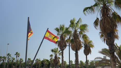 Gente-Caminando-En-Un-Caluroso-Día-De-Verano-Junto-A-La-Playa-De-Valencia,-Paseo-Marítimo-Con-Palmeras-Y-Una-Gran-Bandera-Nacional-De-España