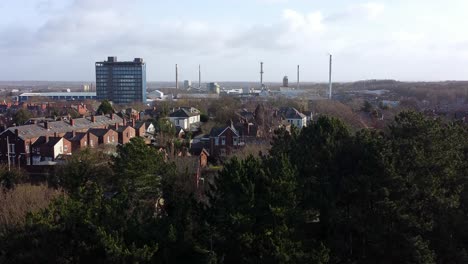Aerial-view-establishing-over-park-trees-to-industrial-townscape-with-blue-Pilkingtons-skyscraper,-Merseyside,-England