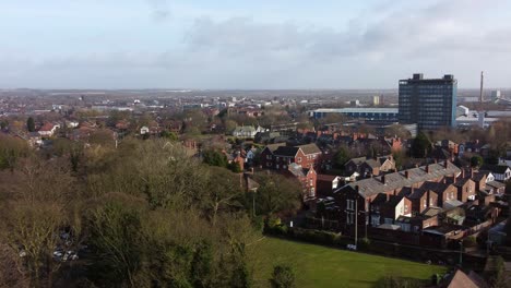Aerial-establishing-view-over-park-trees-to-industrial-townscape-with-blue-skyscraper,-Merseyside,-England