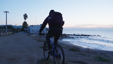 A-traveler-rides-his-bike-towards-a-beach-house,-coming-to-a-stop-at-the-edge-of-the-rocky-shore-to-take-in-the-view-of-the-ocean-during-the-blue-hour