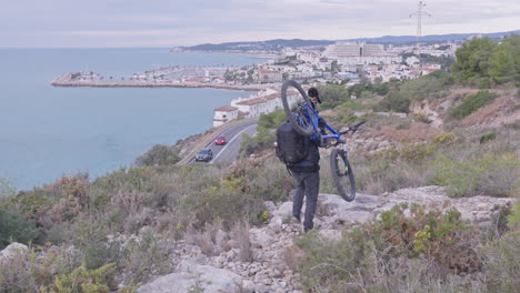 Traveller-carries-his-mountain-bike-on-the-sholder-as-group-of-cyclists-pass-on-a-bendy-road-down-below-leading-to-the-coastal-city
