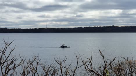 Primary-focus-is-on-foreground-bushes,-with-kayak-passing-by-on-cloudy-day