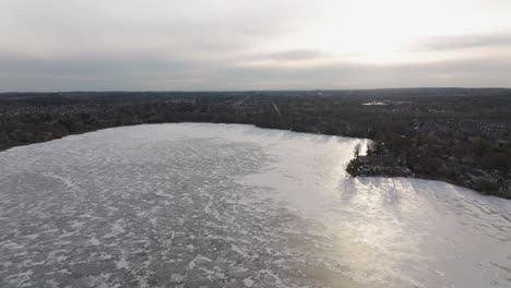 Frozen-Canadina-lake-Wilcox-near-Toronto-in-the-early-spring,-Drone-ascending,-city-panorama