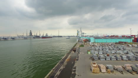 Following-a-ship-with-a-drone-on-the-Scheldt-river-surrounded-by-cargo-ships,-machinery,-granite-blocks,-far-view-from-the-nuclear-station-in-Doel
