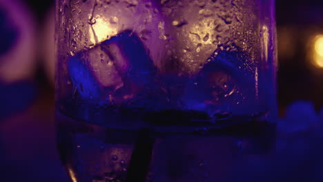 Close-up-of-ice-blocks-falling-into-a-sparkling-drink,-alcoholic-or-non-alcoholic,-surrounded-by-warm-lights-and-night-atmosphere