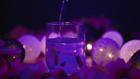Slow-motion-product-video-of-a-lowball-glass-being-filled-with-a-tonic-drink-with-ice-blocks-and-vanilla-sticks,-surrounded-by-atmospheric-lights