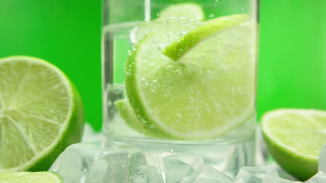 Close-up-product-video-of-a-refreshing-gin-tonic,-alcoholic-summer-drink-with-green-lemons-and-ice-blocks,-isolated-on-a-green-background