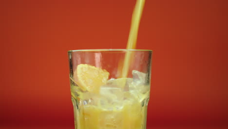 Fresh-orange-juice-pouring-and-spilling-in-a-highball-glass-filled-with-ice-blocks-and-oranges,-isolated-on-an-orange-background