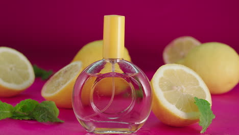 Una-Botella-De-Perfume-De-Vidrio-Con-Una-Tapa-Naranja,-Rodeada-De-Naranjas-Frescas-Y-Un-Fondo-Rosa