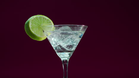 Product-video-of-a-martini-glass,-gin-tonic,-alcoholic-drink-with-a-slice-of-lemon-and-ice-blocks,-rotating-on-a-display-turntable-with-deep-bordeaux-background