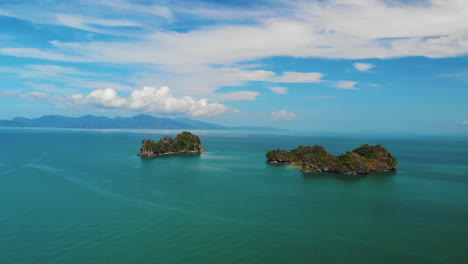 Drone-flying-away-from-two-small-mountainous-islands,-Pulau-Gasing-and-Pulau-Pasir,-shot-from-a-beach-in-Langkawi,-Malaysia