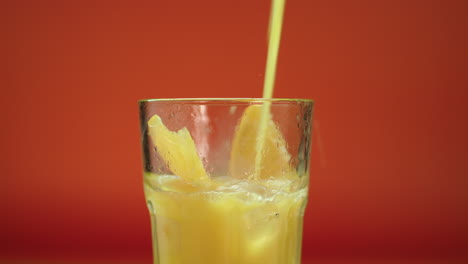 Orange-juice-pouring-in-a-highball-glass-filled-with-ice-blocks-and-fresh-oranges,-isolated-on-an-orange-background