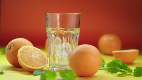 Dropping-ice-blocks-in-an-empty-highball-glass,-surrounded-by-fresh-oranges-and-spearmint,-isolated-on-a-yellow-and-orange-background,-slow-motion-product-video