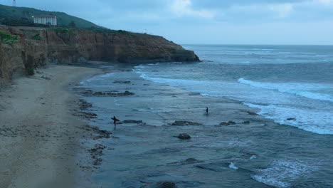 Two-male-surfers-getting-out-of-the-water-at-dusk-at-Sunset-Cliffs,-San-Diego