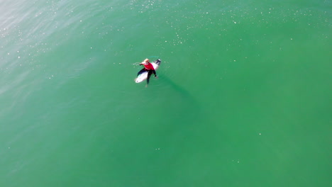Lone-surfer-in-Pacific-Ocean-waiting-for-wave---looking-for-friends,-aerial-4k-drone-boom-down-to-surfer-Zander-Adelsohn-in-red-wetsuit-rash-guard-at-Huntington-Beach,-California-surfing-competition