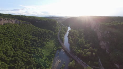 Passing-a-train-on-a-bridge-over-a-river-at-sunset,-surrounded-by-forest---aerial