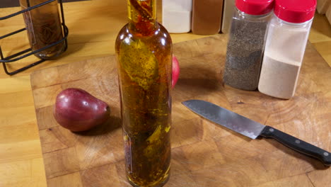 Bottle-of-herb-infused-olive-oil-sits-on-cutting-board-with-knife,-fruit-and-spices