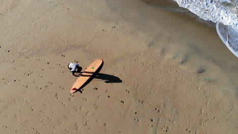 Overhead-aerial-4k-drone-of-female-surfer-girl-walking-with-long-surfboard-and-setting-it-down-in-sand-on-beach-at-Huntington-Beach-Pier,-preparing-to-hit-the-morning-waves