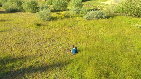 Drone-slowly-circling-a-teenage-boy-sitting-in-the-grass,-being-occupied-with-his-phone
