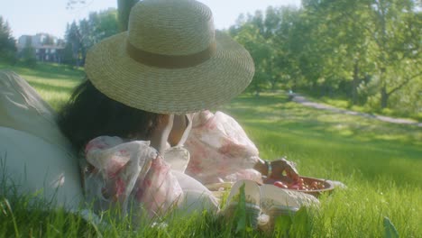 Black-woman-wearing-sun-hat-lying-on-her-side-on-picnic-in-park-eating-grapes