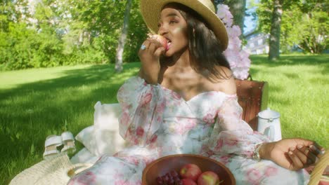 Smiling-black-woman-in-park-on-picnic-in-floral-dress-takes-bite-from-apple
