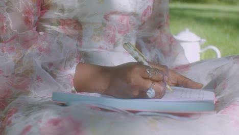 Black-woman-in-white-floral-dress-in-park-writes-in-her-diary,-close-up