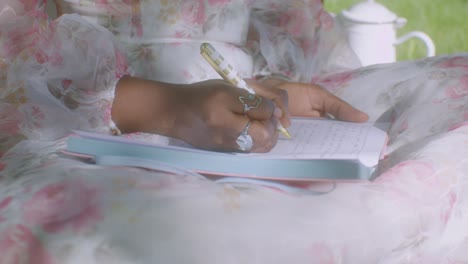 Black-lady-with-floral-dress-sits-down-and-writes-in-diary,-close-up-dolly