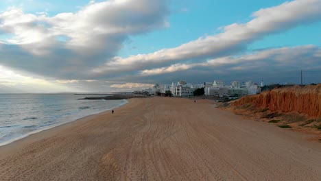 Strand-Mit-Der-Stadt-Quarteira-Im-Hintergrund,-Umgeben-Von-Einer-Klippe,-Einem-Ruhigen-Ozean-Und-Einem-Bewölkten-Himmel