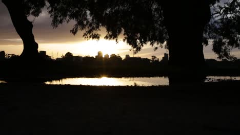 The-sun-sets-from-behind-a-silhouetted-cityscape,-as-a-person-jogs-past-a-pool-of-water-in-the-foreground
