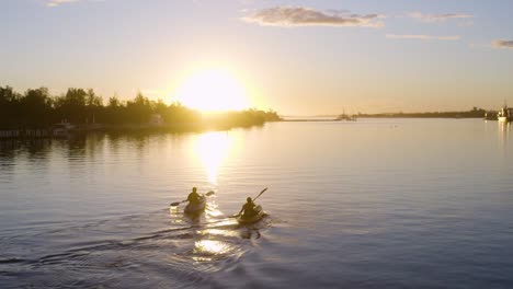 Hermosa-Toma-De-Seguimiento-De-Dos-Kayaks-Navegando-A-Lo-Largo-De-La-Costa-Australiana-Hacia-La-Puesta-De-Sol,-Con-árboles-Y-Cielo-Azul-En-El-Fondo