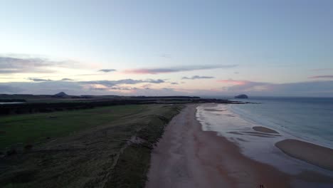Drone-footage-flying-slowly-over-a-long-sandy-beach-and-sand-dunes-during-a-pink-sunset-as-the-tide-gently-laps-the-shore-and-people-walk-along-the-beach