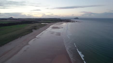 Drone-footage-high-above-a-long-sandy-beach-at-sunset-looking-out-across-the-rippling-ocean-as-the-tide-gently-laps-the-shore