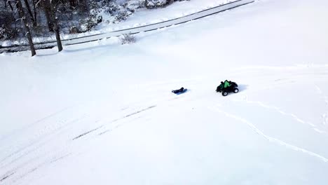 A-drone,-aerial,-birds-eye-top-down-view-of-a-child-riding-on-their-belly-on-a-sled-getting-pulled-by-an-atv-4-wheeler-with-a-child-sitting-on-the-back-in-the-snow-covered-ground-of-the-country