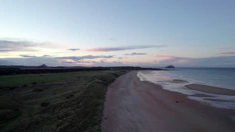 Aerial-drone-footage-rising-up-above-a-long-sandy-beach-and-sand-dunes-during-a-pink-sunset-as-people-walk-along-the-shore
