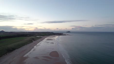 High-panning-drone-footage-flying-above-a-long-sandy-beach-at-sunset-slowly-turning-to-look-out-across-the-rippling-ocean-as-the-tide-gently-laps-the-shore
