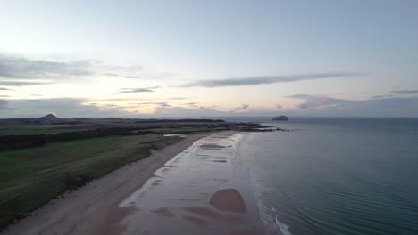 Stationary-drone-footage-flying-high-above-a-long-sandy-beach-at-sunset-looking-out-across-the-rippling-ocean-as-the-tide-gently-laps-the-shore