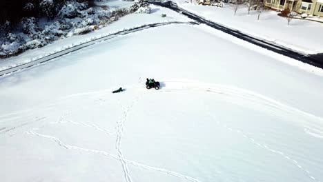 A-drone,-aerial,-birds-eye-view-tracking-an-atv-4-wheeler-with-a-child-riding-on-the-back-and-pulling-another-child-riding-on-their-belly-on-a-winter-sled-on-the-snow-covered-ground-in-the-country