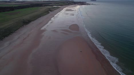 Drone-footage-high-above-a-long-sandy-beach-tilting-to-reveal-people-walking-along-the-beach-and-a-sunset,-looking-out-across-the-ocean-as-the-tide-gently-laps-the-shore