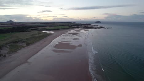 Fast-aerial-drone-footage-flying-high-above-a-long-sandy-beach-at-sunset-looking-out-across-the-rippling-ocean-as-the-tide-gently-laps-the-shore