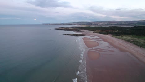 Slow-moving-drone-footage-flying-towards-the-rising-moon-high-above-a-sandy-beach-and-ocean-while-the-tide-laps-the-shore