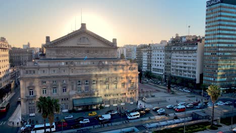 Aerial-panning-view-capturing-colon-theater-on-the-rear-entrance,-busy-downtown-rush-hour-traffics-on-9-de-julio-avenue-with-big-glowing-sun-in-the-background-at-sunset,-Buenos-Aires-city,-Argentina