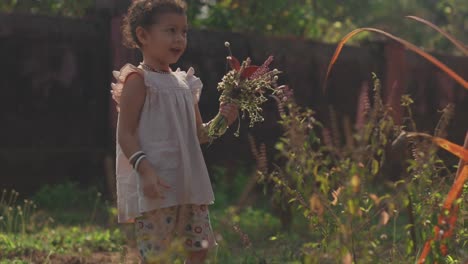 Ein-Hübsches-Kleines-Mädchen,-Das-Einen-Strauß-Wildblumen-Sammelt,-Während-Sie-Draußen-In-Der-Sonne-Durch-Einen-Garten-Geht-Und-Blumen-Pflückt